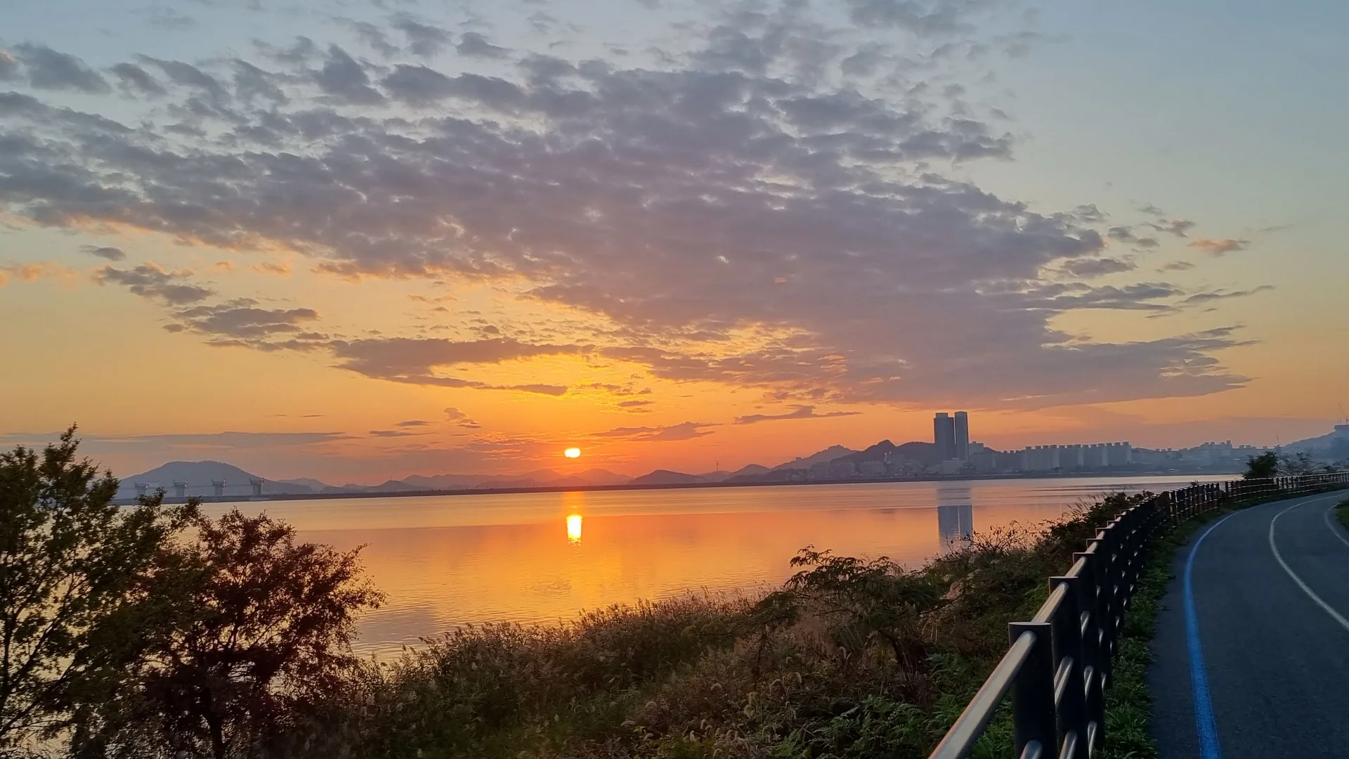 A flat riverside bike path at sunset, the blue route line running ahead with the water on the left