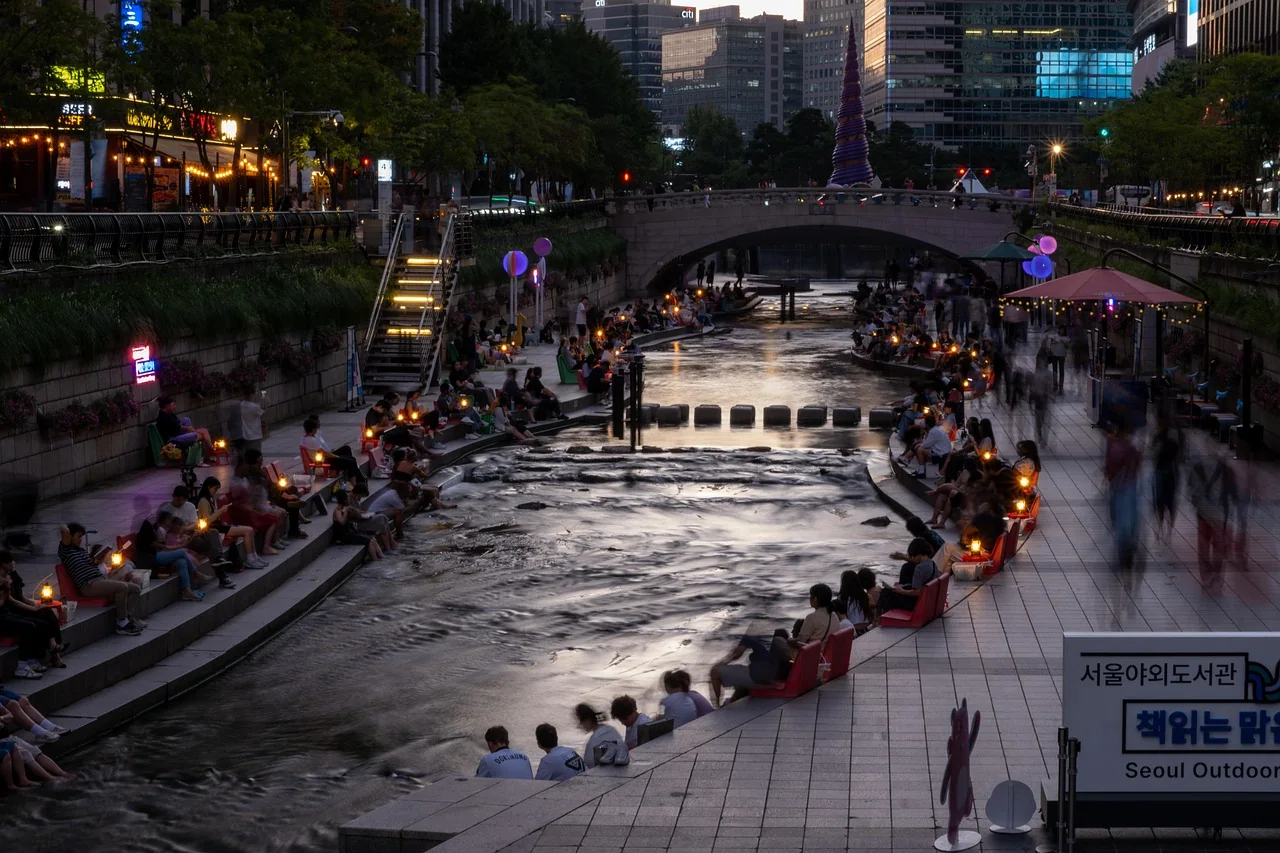 People sitting along a restored stream below city towers at dusk