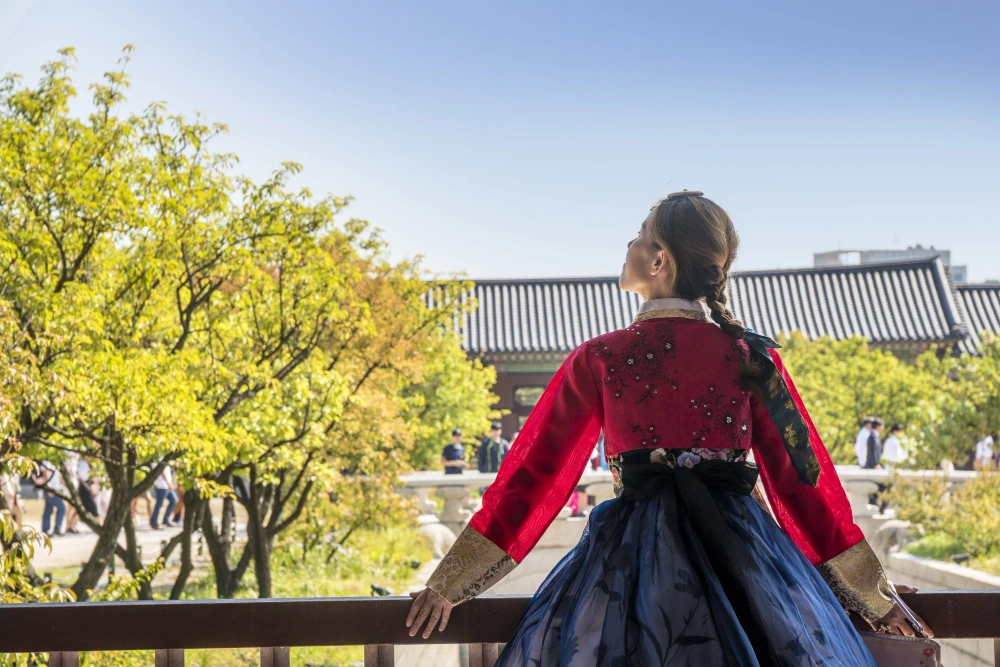 Visitor in hanbok looking out over a palace roofline from a raised walkway