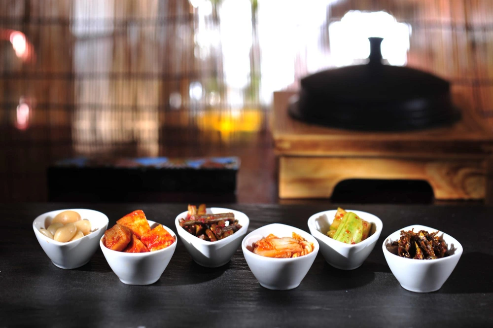 Six small bowls of side dishes lined up on a dark counter