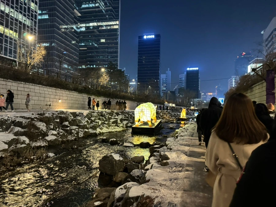 Glowing animal sculpture standing in a shallow stream with onlookers on the walkway above