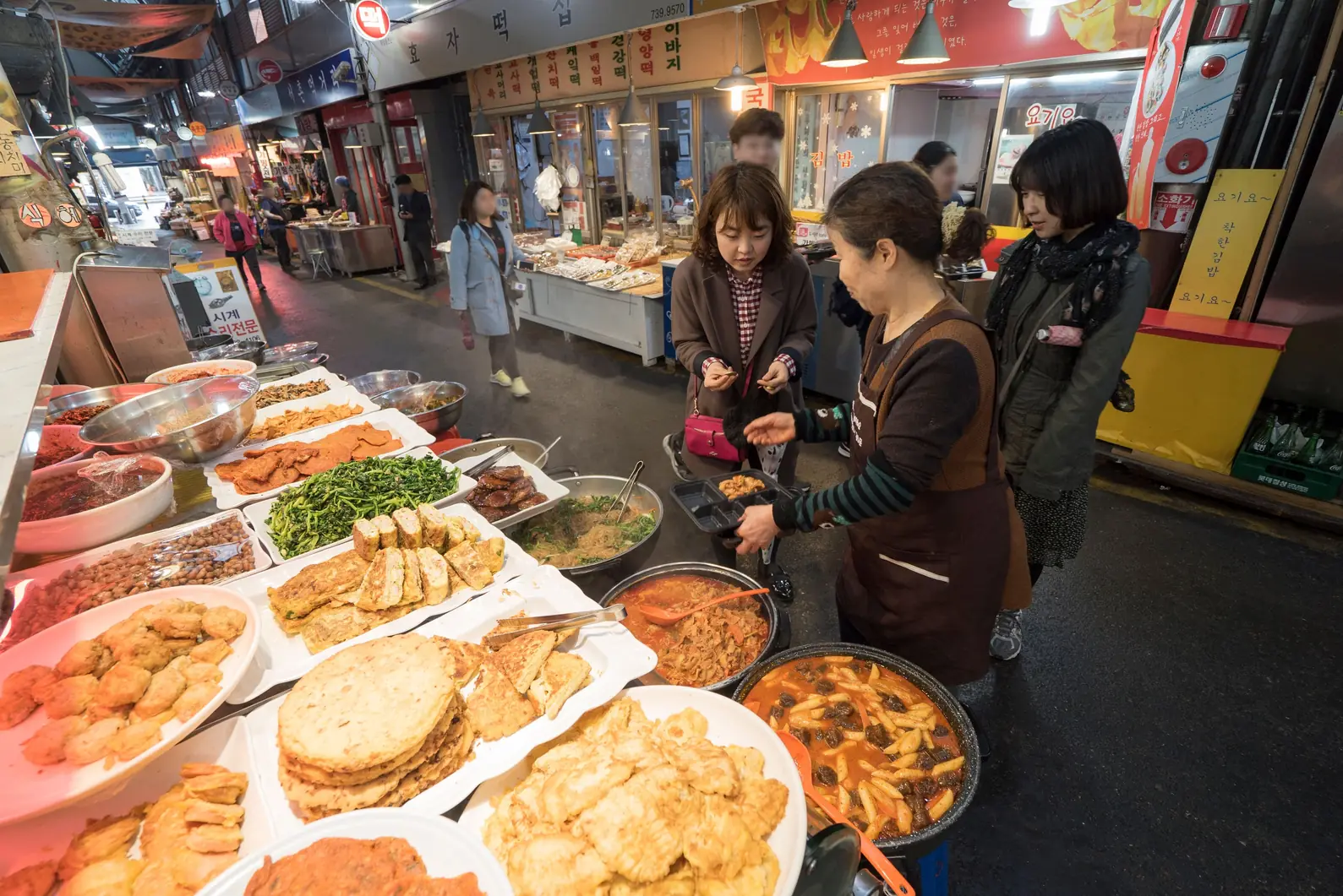 Market vendor handing a lunch tray across a counter of side dishes to waiting customers