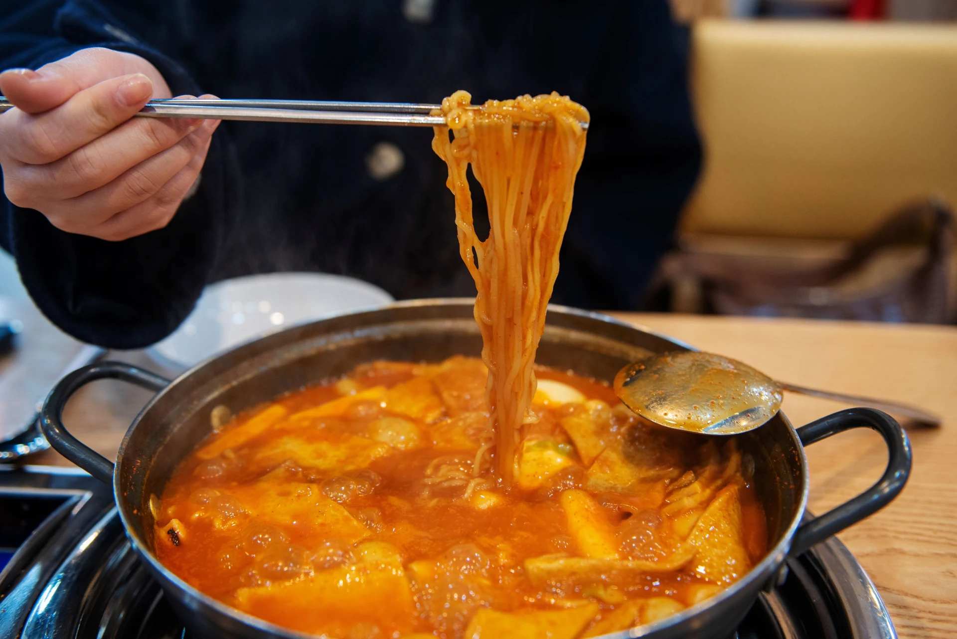 Chopsticks lifting noodles from a pan of red rice cakes and broth