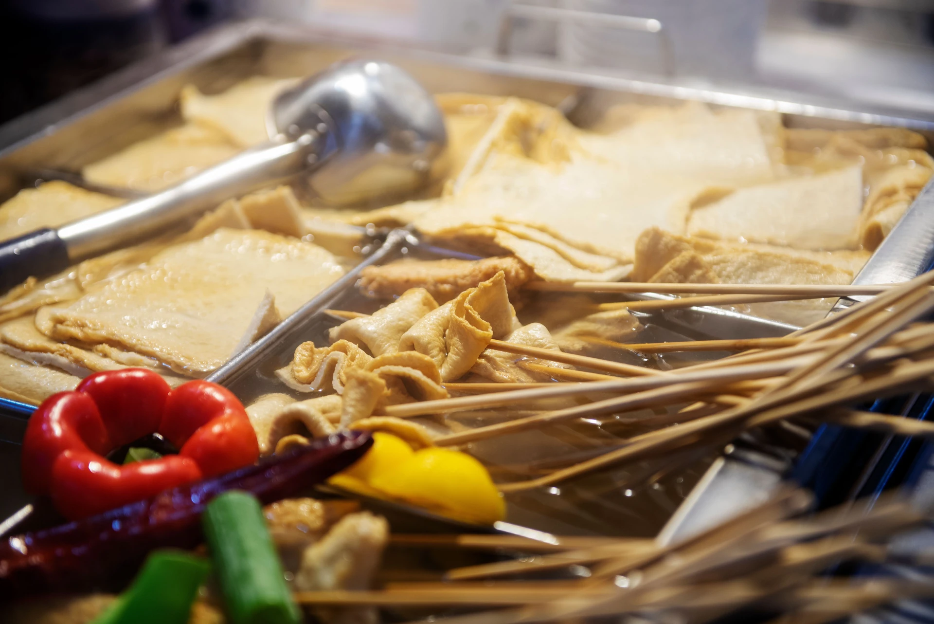Tongs and a ladle resting in a pan of fish cakes in broth beside a bundle of skewers