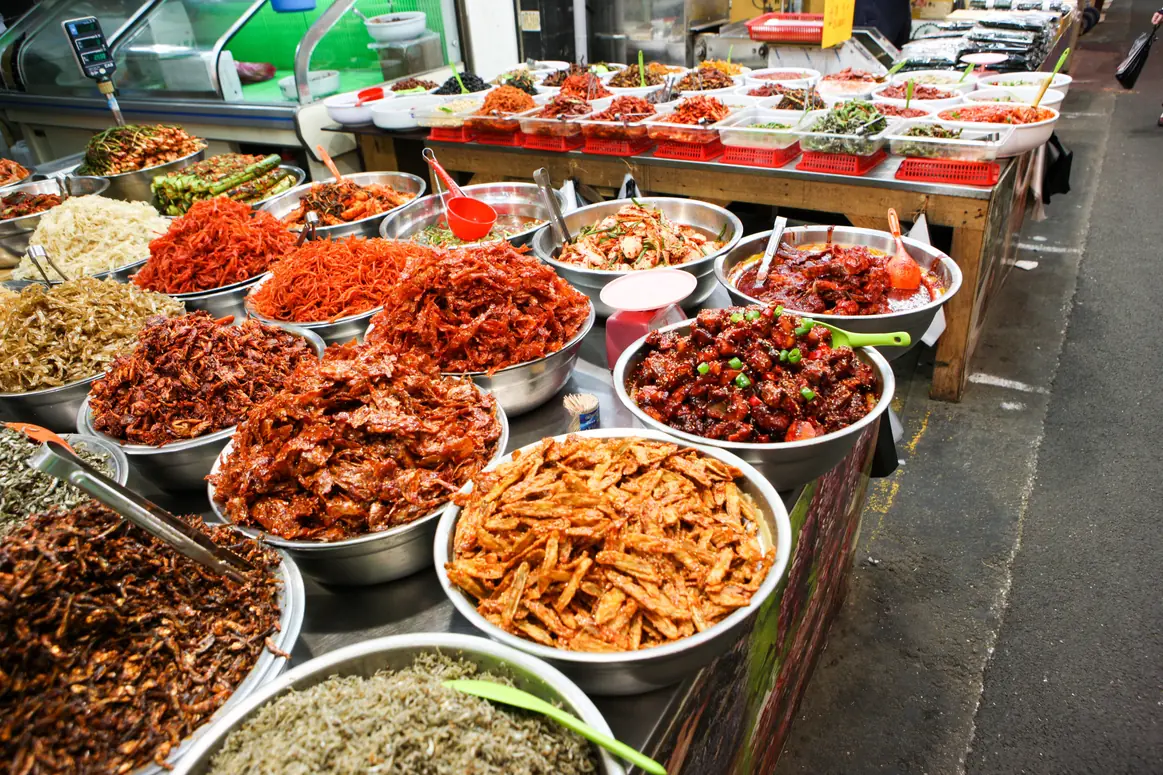 Fifteen steel basins of prepared side dishes along a stall counter with tongs and a scale