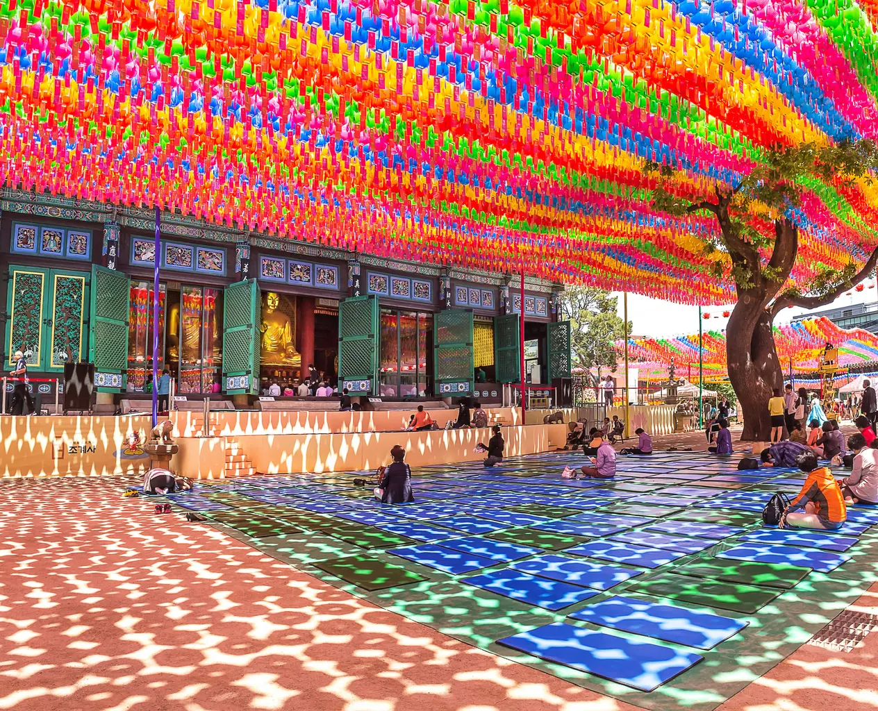 Rows of coloured paper lanterns strung above a temple courtyard where visitors sit on mats