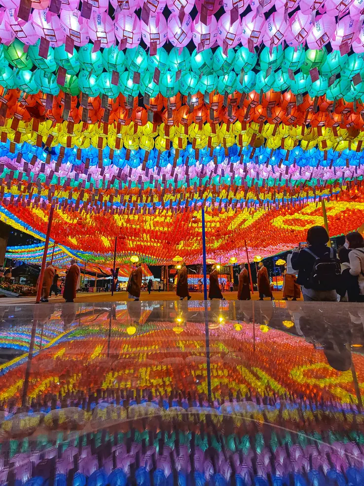Monks in robes lined up beneath a dense canopy of coloured lanterns, reflected on a wet floor
