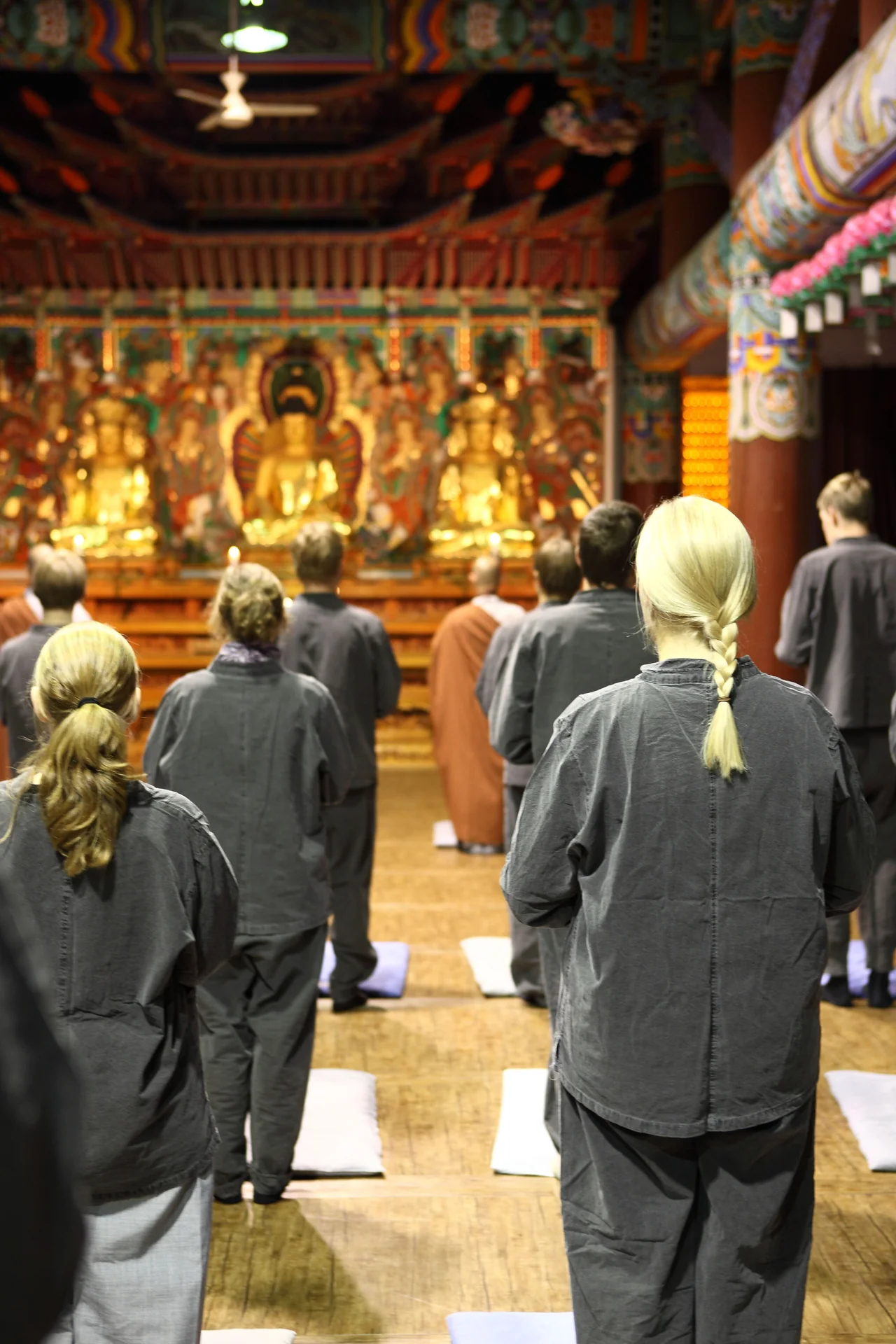 People in grey temple clothing standing in rows facing gilded statues, with a robed monk among them