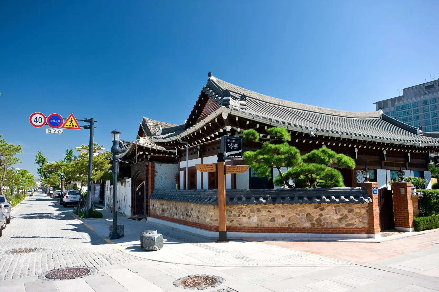A tiled-roof wooden building with pine trees at a street corner, road signs at the left