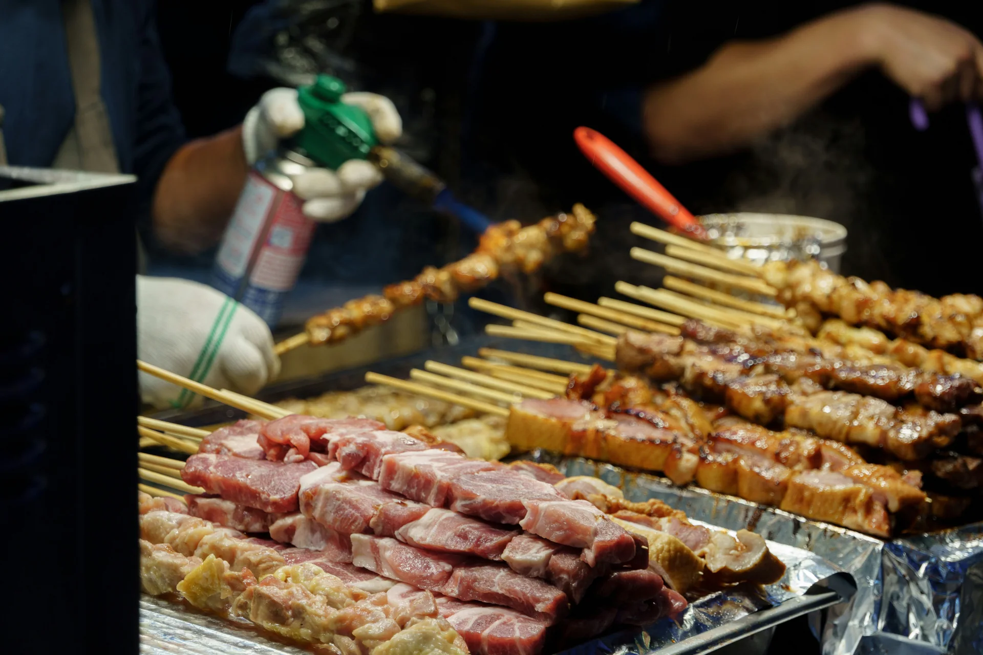 Vendor searing skewers with a hand torch above a tray of raw cuts