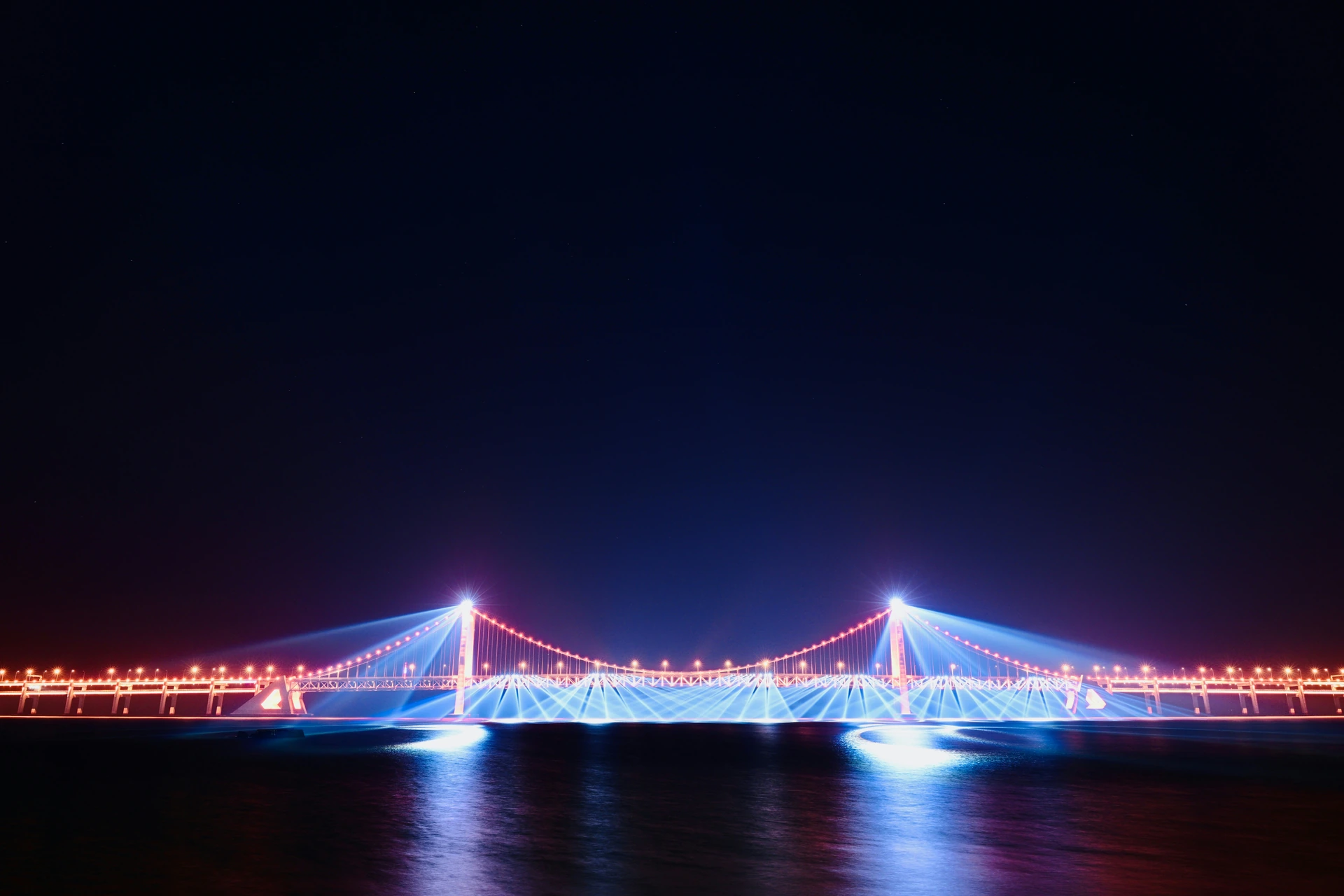 Lit suspension bridge reflected across dark water at night