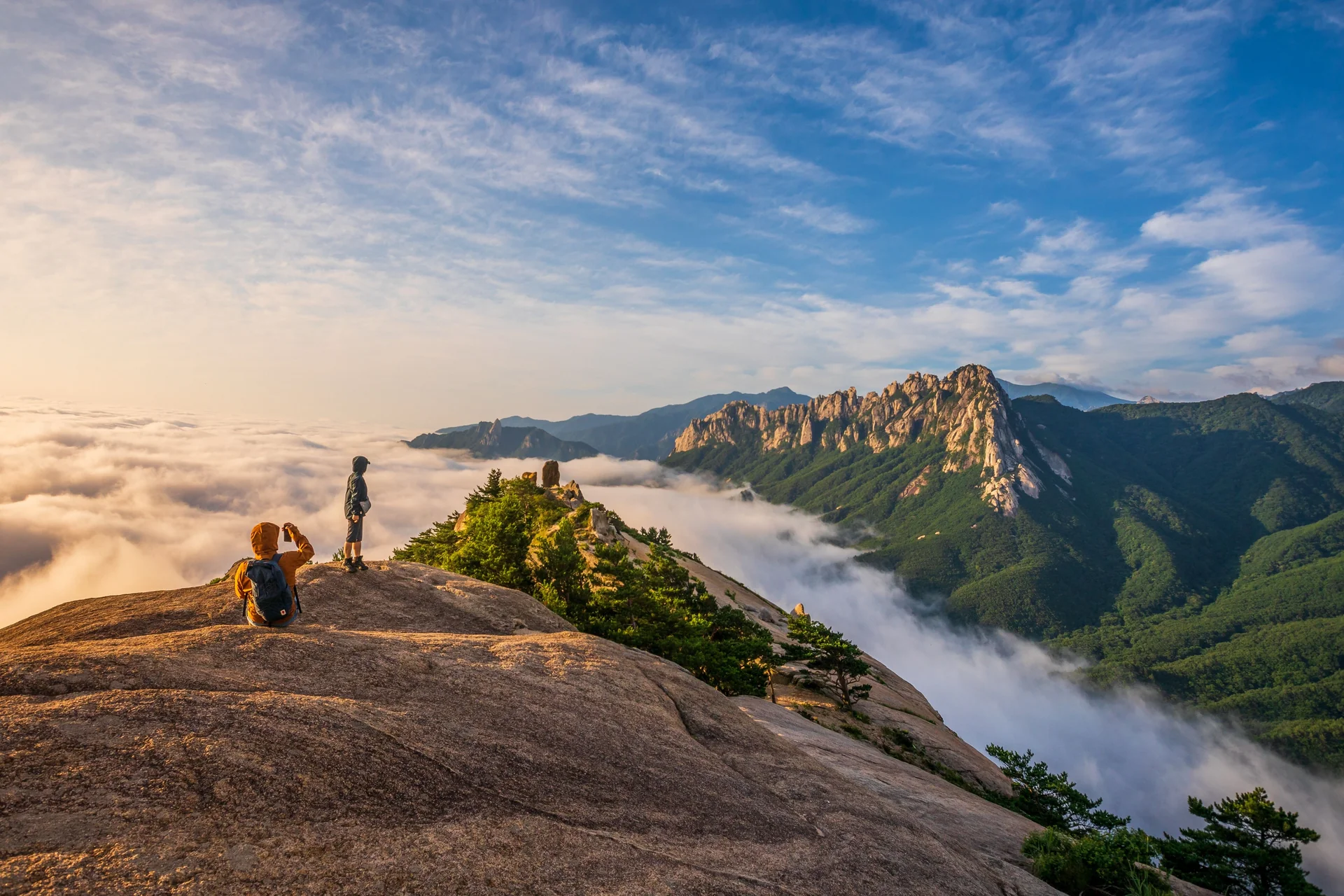 Two hikers on a granite summit above a sea of cloud, with a jagged ridge and forested slopes beyond