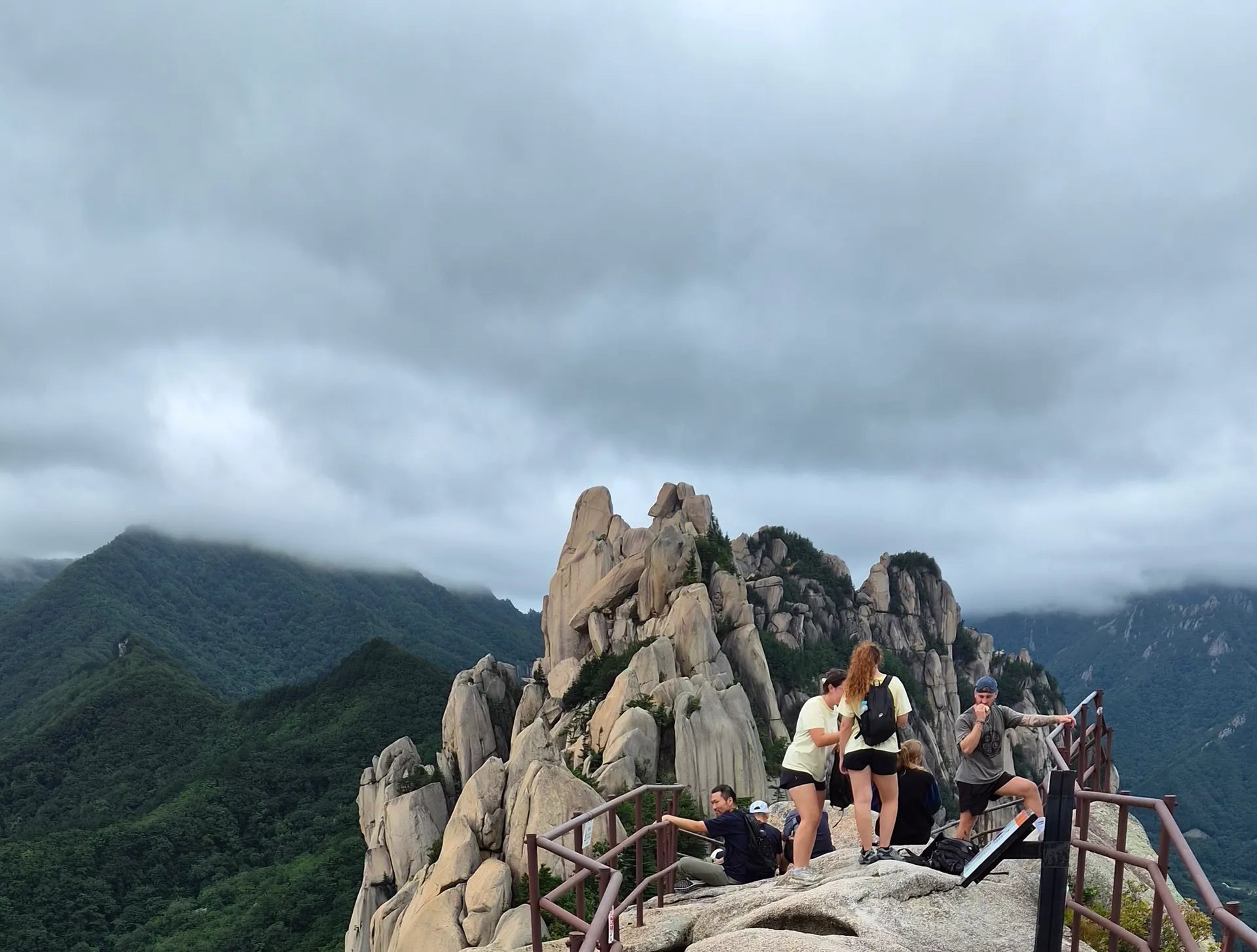 Walkers resting on a rock summit with railed paths and cloud over the ridges behind