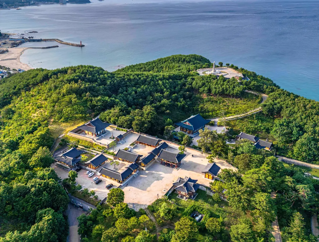 Tiled temple halls on a wooded headland above the East Sea, with a beach and harbour along the shore