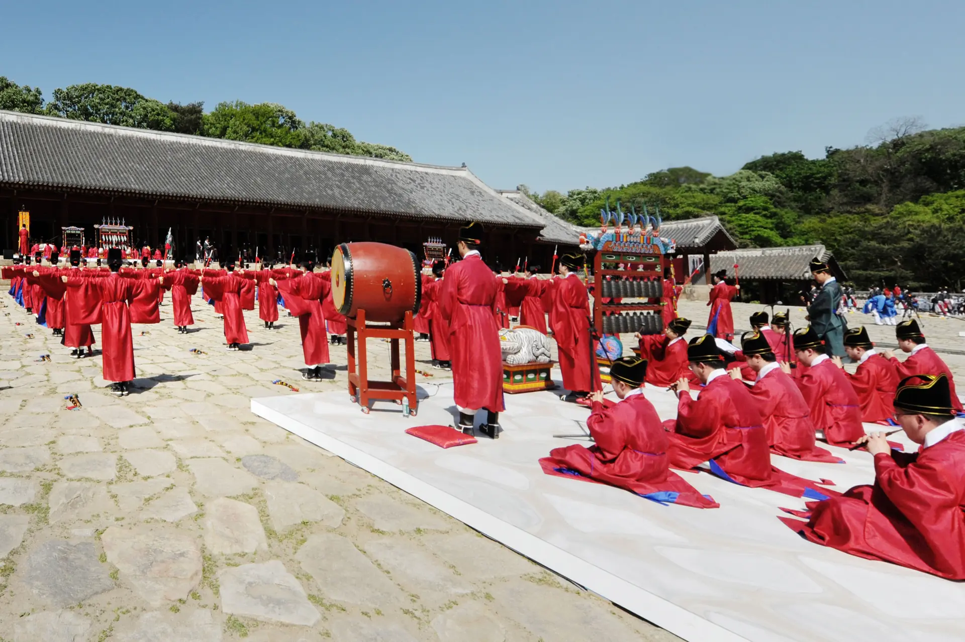 Dozens of people in red ceremonial robes with a large drum on a stone courtyard