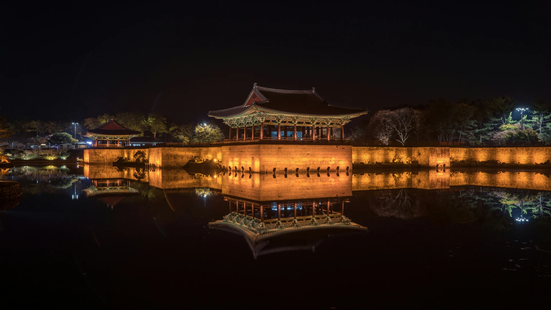 A lit wooden hall and pavilions reflected in still water at night